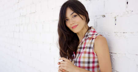Woman leaning against brick wall painted white fiddles with her long brown hair and stares at the cameraの写真素材
