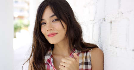 Beautiful woman in long hair smiling with hand up near collar while standing beside white wallの写真素材