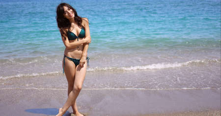 Pretty sexy young woman in a bikini standing at the edge of the surf on a tropical beach smiling at the cameraの写真素材