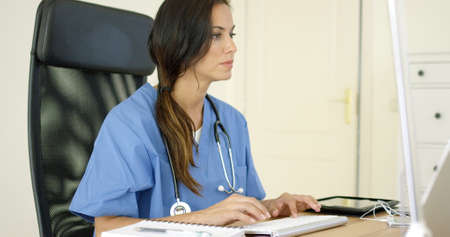 Young female doctor sitting typing in her office as she enters patient records onto a central...