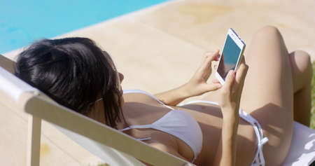 Young woman browsing on a mobile in the summer sun as she relaxes in her bikini on a deck chair near...