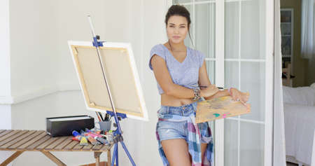 Sexy attractive young female artist standing working on a canvas on an easel on an outdoor patio in...