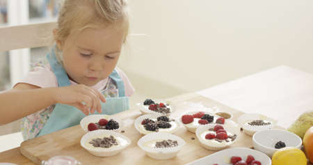 Adorable little blond girl with blond hair carefully putting berries in muffin cups ready to be bakedの写真素材