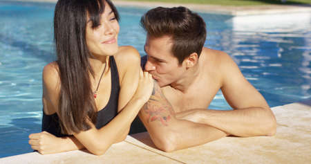 Man in pool standing with girlfriend kisses her hand as the sun shines down on themの写真素材