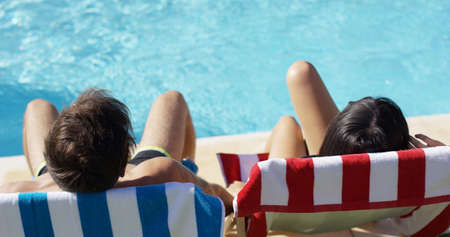 Couple relaxing in colorful deck chairs at the pool enjoying the hot summer sun  view from the rear with just their head visibleの写真素材