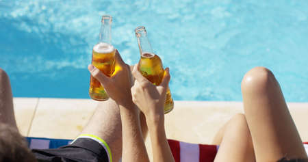Couple relaxing at the pool with beers enjoying their summer vacation  close up view of their hands and the full bottlesの写真素材