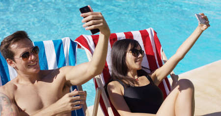 Man and woman taking self portraits with their smart phones at while seated in deck chairs at the swimming pool outsideの写真素材