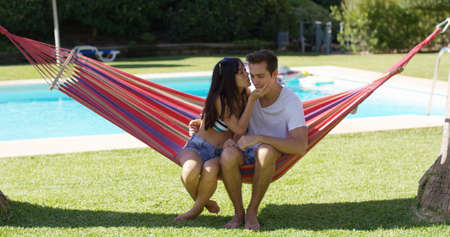 Loving couple in hammock kissing each other with swimming pool behind them during summerの写真素材
