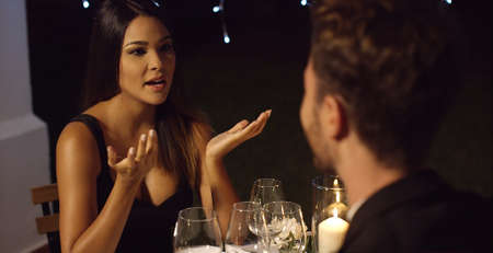 Elegant young woman enjoying a dinner date talking to a young man opposite her at the restaurant table gesturing with her handsの写真素材