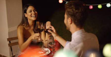 Couple laughs as they raise their red wine glasses while on a night time date in fancy outdoor restaurant on Valentines dayの写真素材