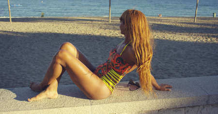 Young woman with lovely long hair relaxing at the seaside in a colorful swimsuit sitting on a wall overlooking a beach and calm ocean looking away from the camera.の写真素材