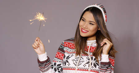 Fun young woman celebrating Christmas with a sparkler watching the fiery sparks with a happy grin in a stylish winter outfitの写真素材