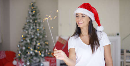 Joyful young woman celebrating Christmas at home holding a burning sparkler in her festive red Santa hat in front of a decorated treeの写真素材
