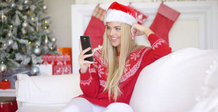 Young happy blond girl in Christmas outfit using mobile phone while sitting on sofa in beautiful...