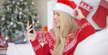 Young happy blond girl in Christmas outfit using mobile phone while sitting on sofa in beautiful...