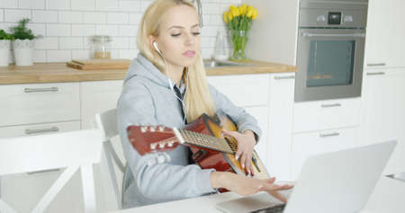 Young wonderful female sitting at table and using laptop with headphones while playing guitar in kitchen alone.の写真素材