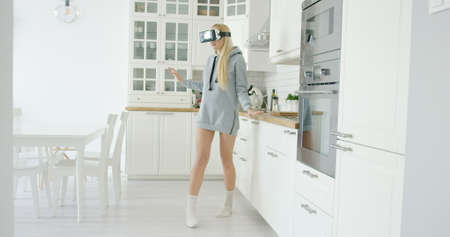 Young girl in grey sweat shirt wearing VR glasses and standing among light and modern kitchen alone.
