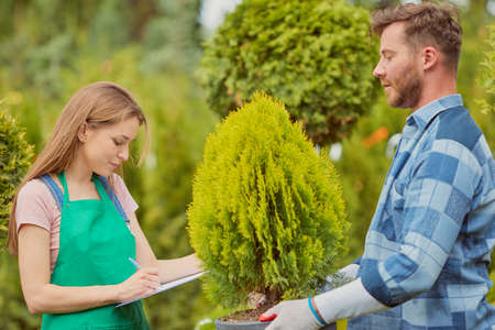 Young gardener woman standing and writing in documents while man doing horticulture job in the...