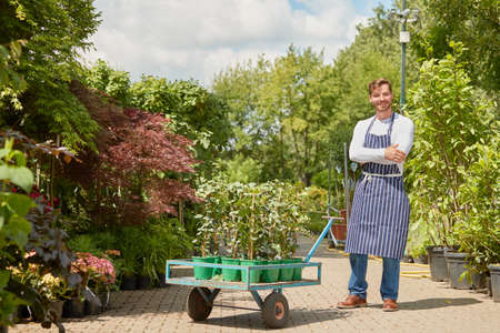 Horizontal outdoors shot of smiling male gardener standing with wagon with potted plants and looking at camera.の写真素材