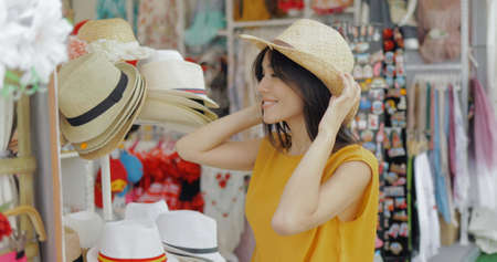 Young cheerful girl posing in shop of accessories trying on straw hat and looking at mirror while shopping in mall.の写真素材