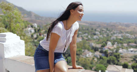 Pretty brunette in casual outfit looking excited while posing on viewpoint and exploring coastal city from height in summertime.の写真素材