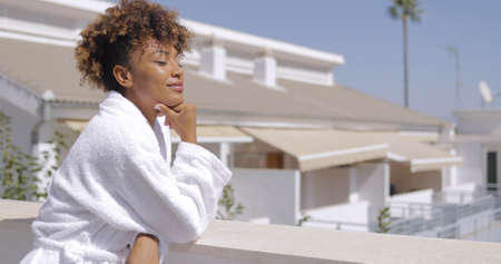Young gentle girl with curls posing in white bathrobe on terrace of resort hotel leaning on fence with eyes closed and enjoying morning sunshine.の写真素材