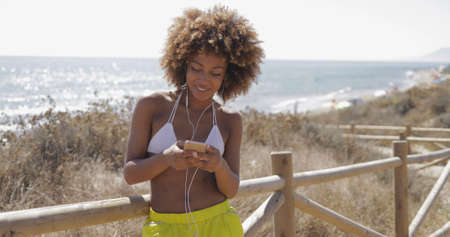 Beautiful curly girl in swimsuit and sportive shorts posing cheerfully with smartphone in hands using it and listening to music with headphones.の写真素材