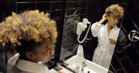 Young ethnic woman in white soft bathrobe looking in glass doing hairstyle while preparing inside of hotel bathroom.の写真素材
