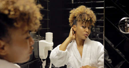 Attractive young woman in white soft bathrobe looking in mirror while posing in modern bathroom of hotel and preparing.の写真素材