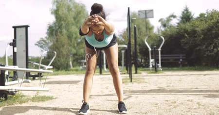 Anonymous ethnic woman in sportswear bending forward and stretching body while standing on sports...