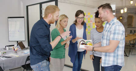 Side view of young man holding box with doughnuts and treating delighted coworkers gathering in...