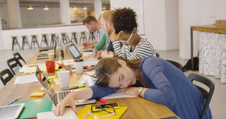 Young exhausted female employee lying on table and sleeping on background of working colleagues.の写真素材