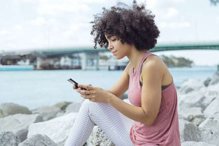 Pretty black woman sitting on rocky river shore with city on background and surfing smartphone in...