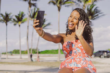Cheerful African-American woman in dress waving at camera of smartphone while having videocall on background of summer park.の写真素材