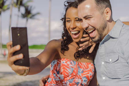 Cheerful multiracial man and woman using smartphone and taking selfie in bright sunlight on background of tropical park.の写真素材