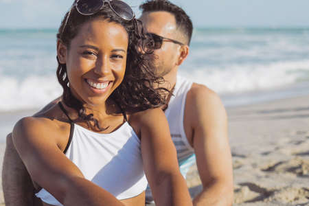 Wonderful laughing African-American girl in bikini sitting with man on sandy beach in tropical sunlight and smiling at camera.の写真素材