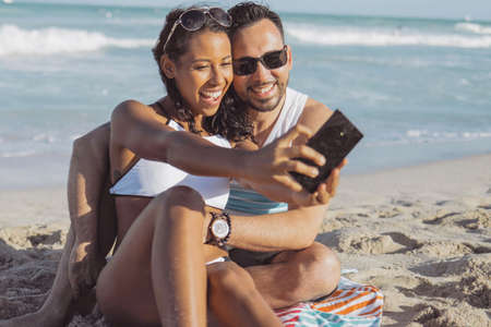 Content black girl with man sitting on sandy beach and using camera of smartphone and taking selfie for memory.の写真素材