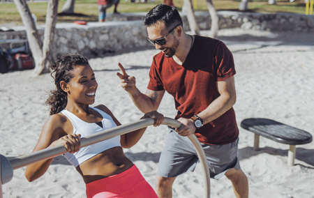 Cheerful man supporting young black woman while doing chin-ups on sandy beach with gym constructions.の写真素材