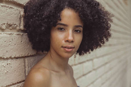 Headshot of young African-American girl with nude shoulders and short rich curls looking at camera leaning on wall.の写真素材