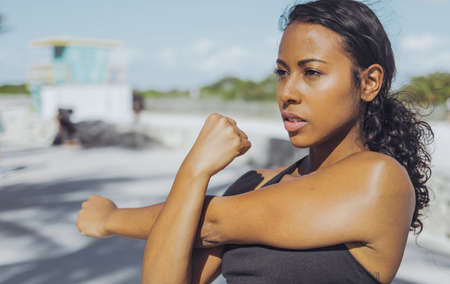 Confident African-American woman in sportswear training on waterfront in sunlight warming up hands and looking forward.の写真素材