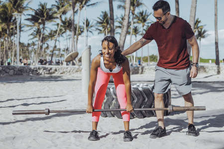 Serious sportive man supporting black woman doing deadlift with metal barbell standing on beach gym in sunlight.の写真素材