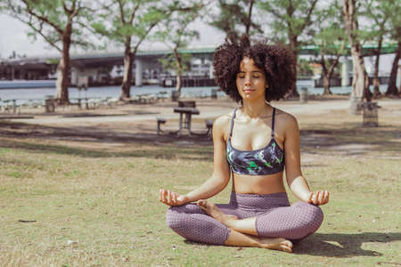 Relaxed fit black girl in sportswear sitting with legs crossed on green meadow in park and meditating with eyes closed.の写真素材