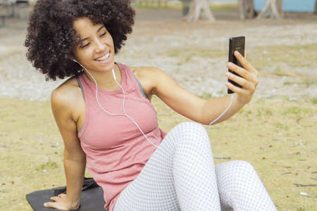 Pretty African-American girl in sportswear sitting on mat in park and chatting online wearing headphones and using phone.の写真素材