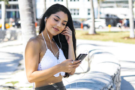 Casual young woman with long beautiful hair sitting on fence of tropical alley and listening to music with headphones and phone.の写真素材