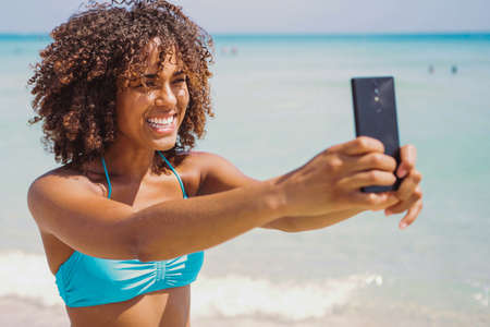 Cheerful young pretty ethnic woman standing on the beach and taking selfie with smartphone at...