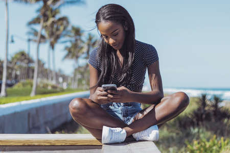 Beautiful young black woman sitting and using smartphone on fence in sunny summer day on the background of the sea.の写真素材