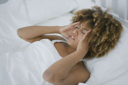 Beautiful unhappy woman lying in bed under blanket and suffering from awful headache closing eyes and touching temples.の写真素材