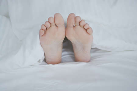 Crop shot of woman lying under cozy white blanket in bed sticking out feet while sleeping and relaxing.の写真素材