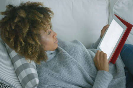 Ethnic young model lounging on comfortable sofa and chilling while browsing tablet in morning time.