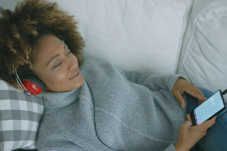 Content young model holding smartphone and wearing headphones relaxing on sofa and listening to...